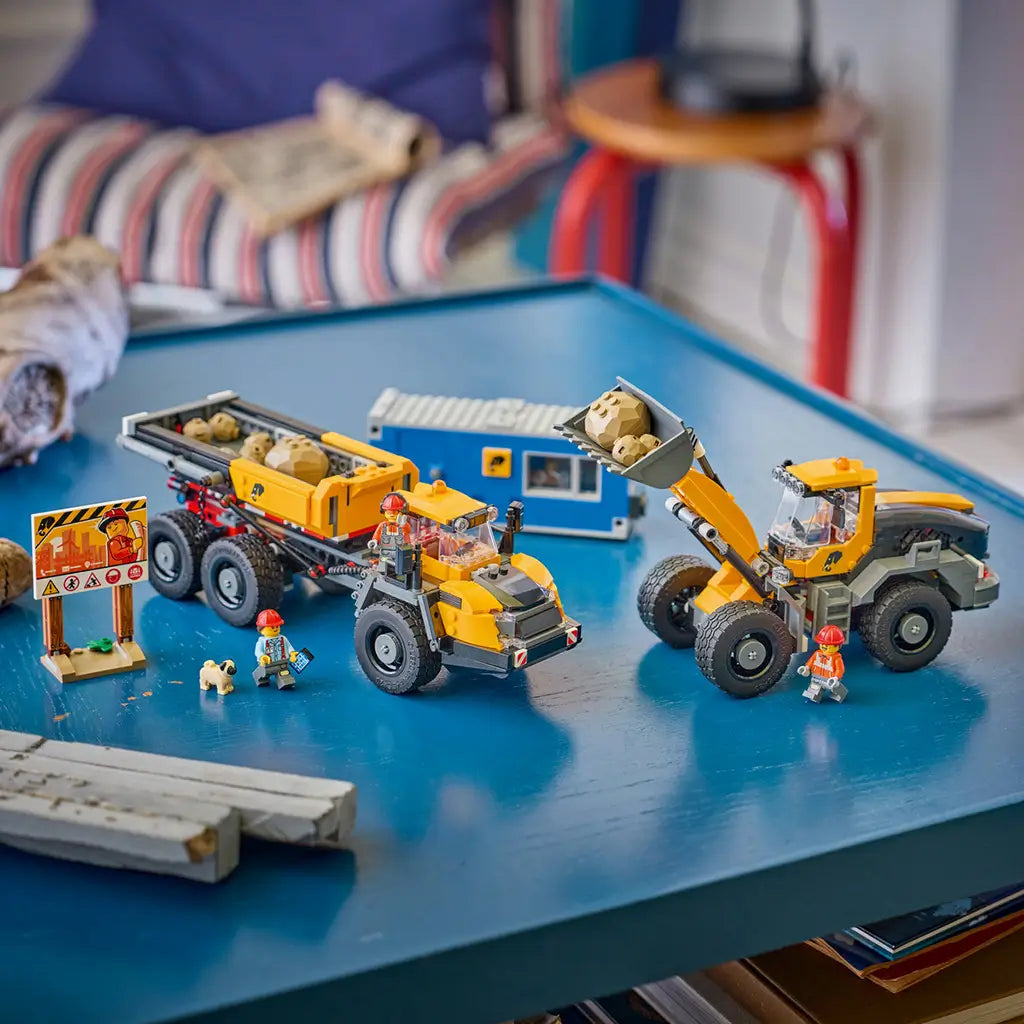 LEGO City Dump Truck & Front End Loader in yellow, black, gray, and blue plastic bricks is displayed on a blue table with workers, tan rocks, a site office, safety sign, and dog.