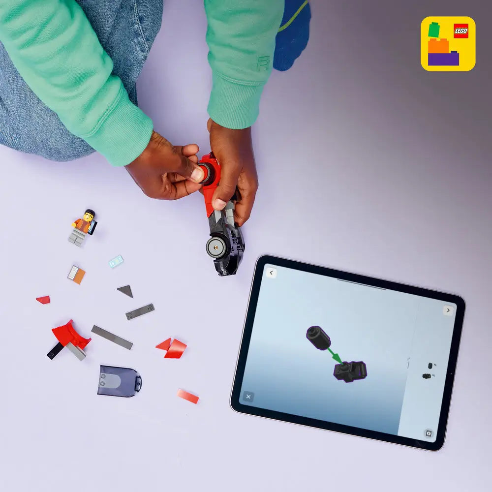 LEGO City EV Supercar building process with black and red plastic pieces, angular curved parts, and small wheels, assembled by hands beside a tablet showing digital instructions on a light tabletop workspace.