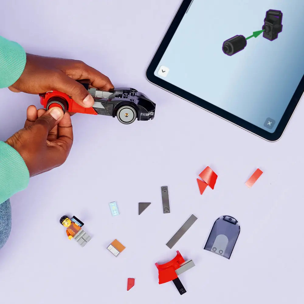 LEGO City EV Supercar partially assembled in black and red plastic bricks, compact low-profile shape, with loose pieces scattered on a light surface as hands follow digital building instructions on a tablet.