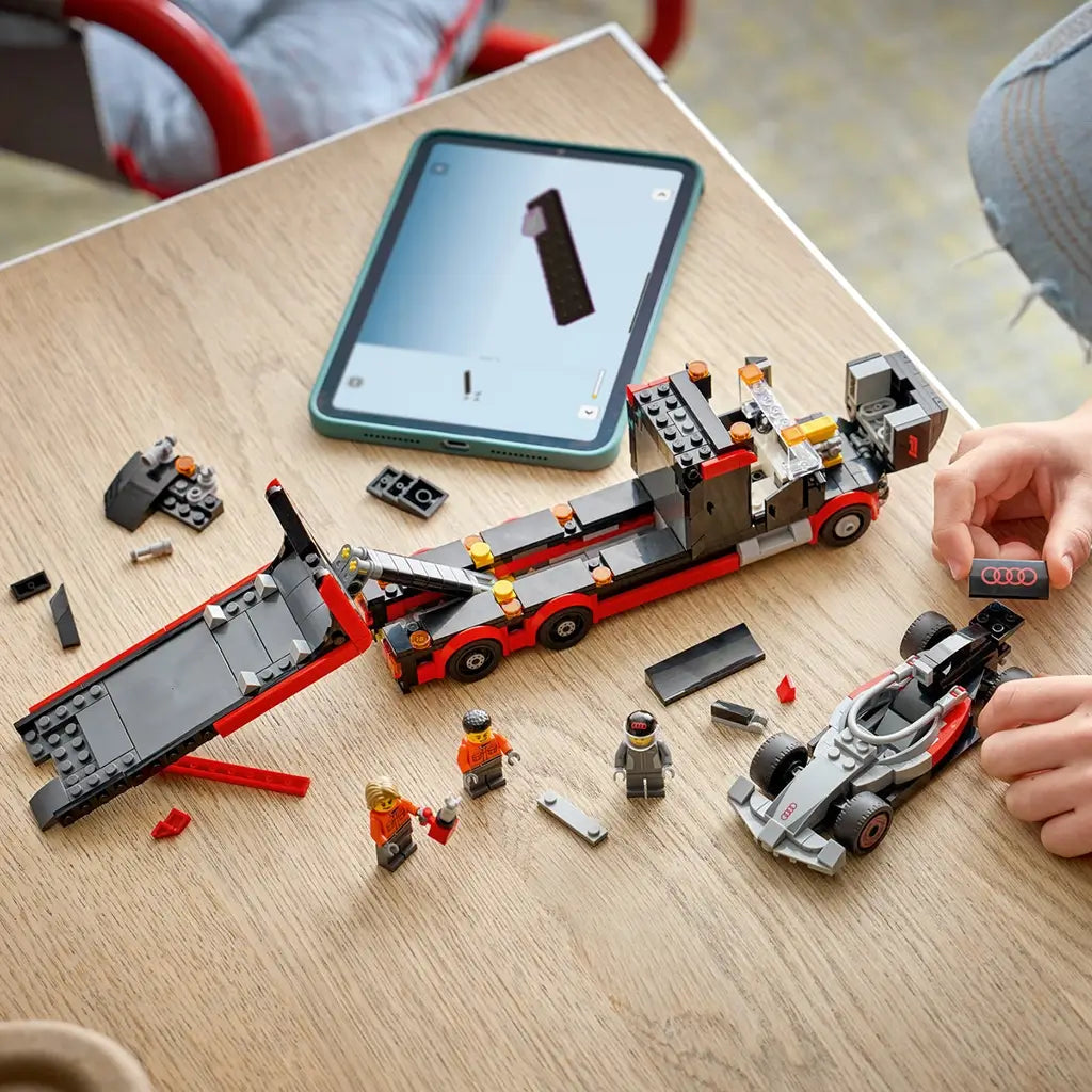 LEGO City F1 truck build in red, black, and gray plastic pieces, flatbed vehicle and race car components, construction play scene on a wooden table with tablet instructions in background.