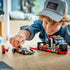 LEGO City race car playset in red, black, and gray plastic, flatbed truck and Formula 1 car models, child assembling toy vehicles on a wooden table in a bright indoor setting.