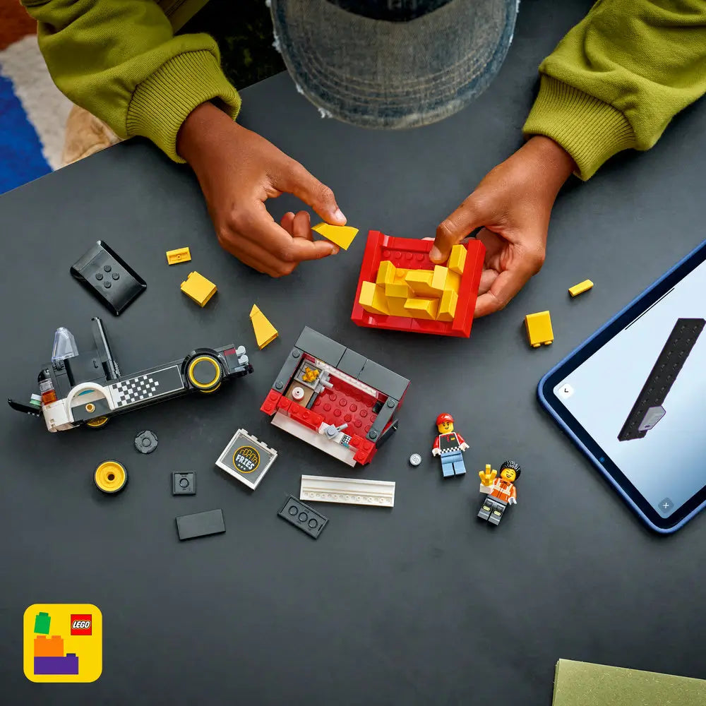 LEGO City fries food truck build scene showing hands assembling a red fry container roof detail from yellow bricks, with scattered parts, minifigures, and digital instructions on a tablet nearby.