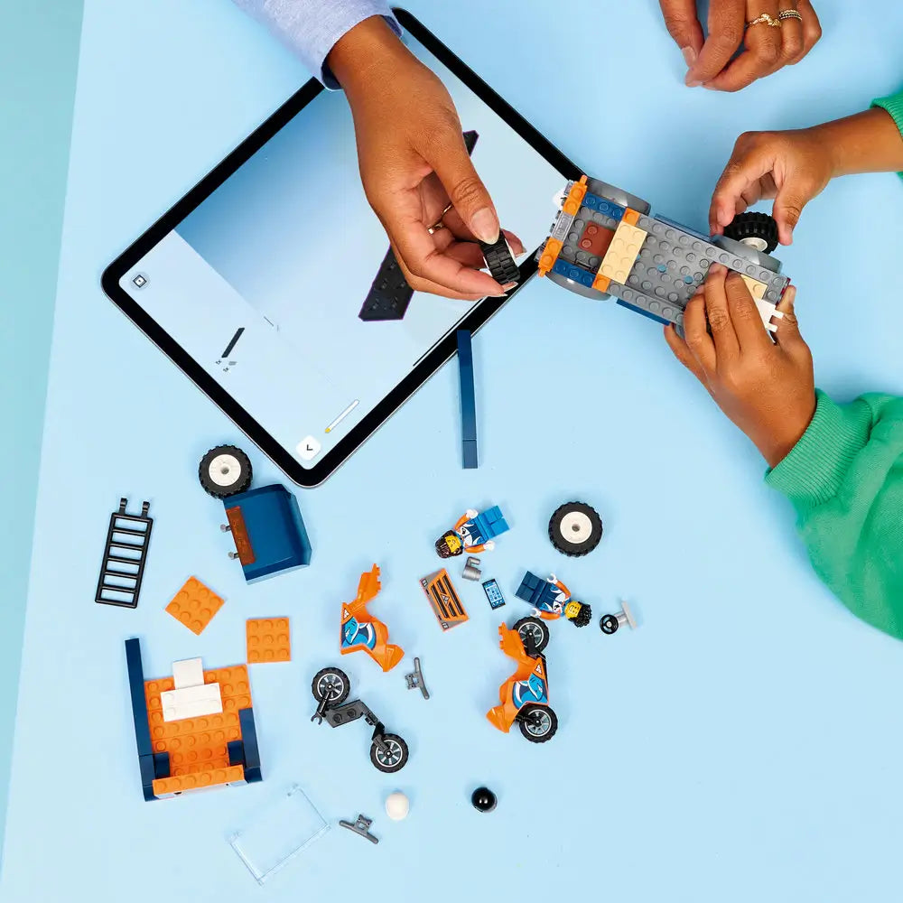 LEGO City motorcycle transporter building set shown during assembly, featuring blue and orange truck parts, loose wheels, motorcycle pieces, minifigures, and digital instructions on a tablet.