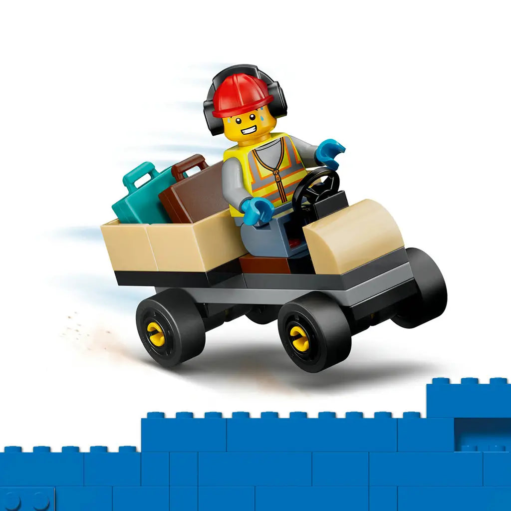 LEGO City airport worker minifigure wears a red helmet and gray safety vest while driving a tan plastic baggage cart loaded with luggage on a white background.