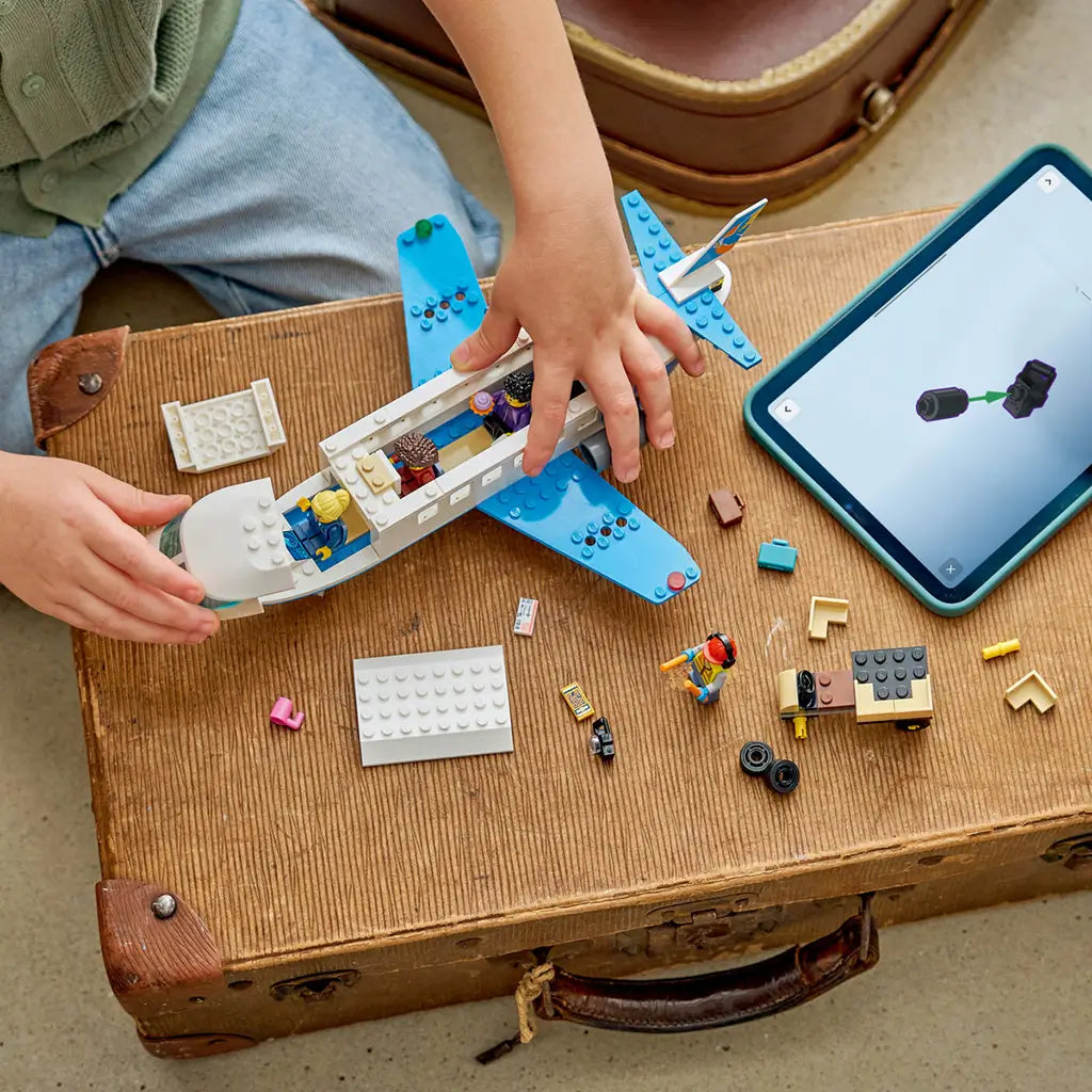 LEGO City Passenger Jet building toy in white and blue plastic is assembled by a child on a vintage wood suitcase beside loose bricks, minifigures and a tablet.