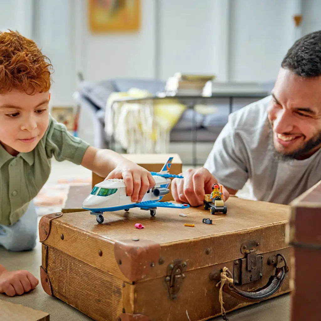 LEGO City Passenger Jet toy in white and blue plastic is played with by a child and adult beside a baggage cart on a vintage brown suitcase in a bright living room.