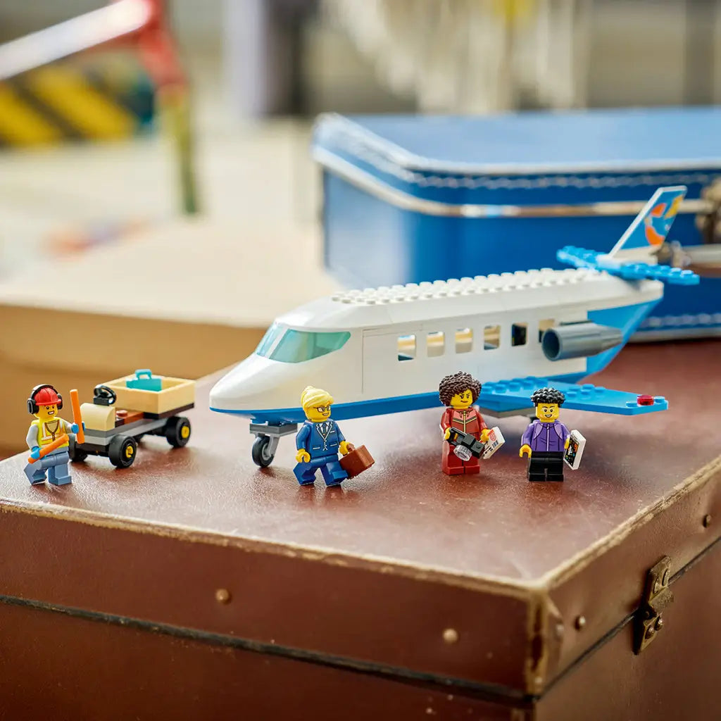 LEGO City Passenger Jet display features a white and blue plastic airplane, four minifigures and a tan baggage cart arranged on a brown vintage suitcase indoors.