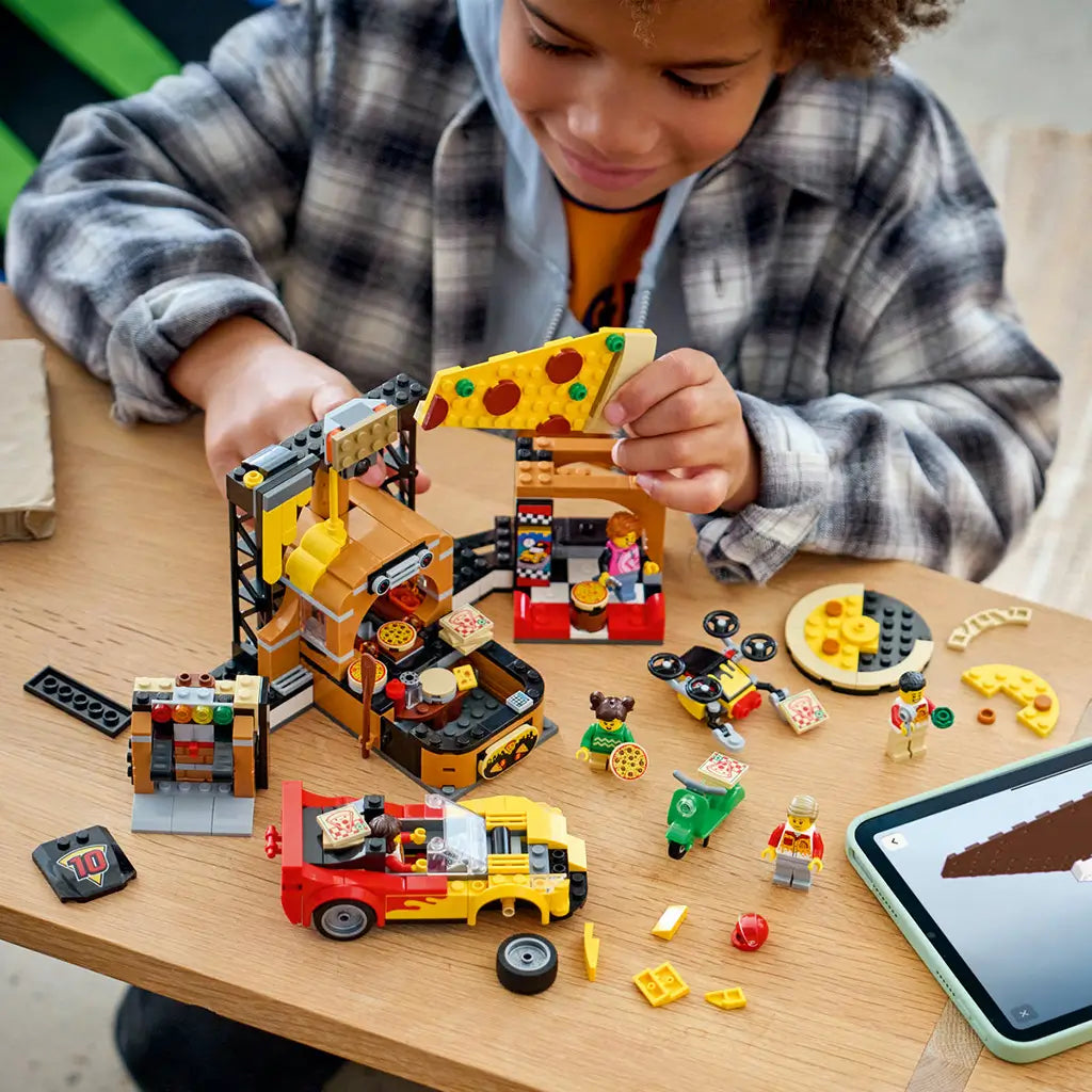 LEGO City Pizza Delivery Experience in yellow, brown, red, and green plastic bricks is assembled by a child at a table with a pizza shop, car, scooter, drone, and loose pieces.