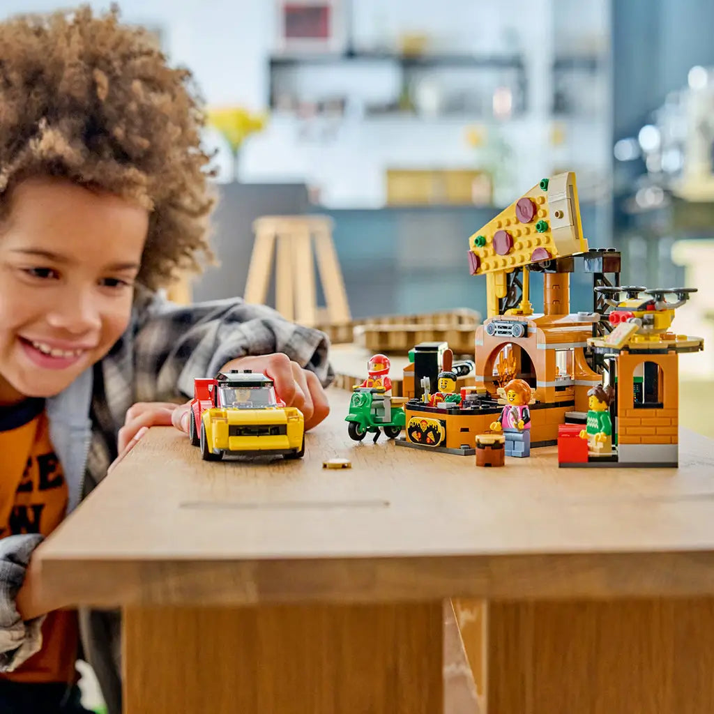 LEGO City Pizza Delivery Experience in yellow, brown, red, and green plastic bricks is played with by a smiling child, with a pizza shop, sports car, scooter, and minifigures.