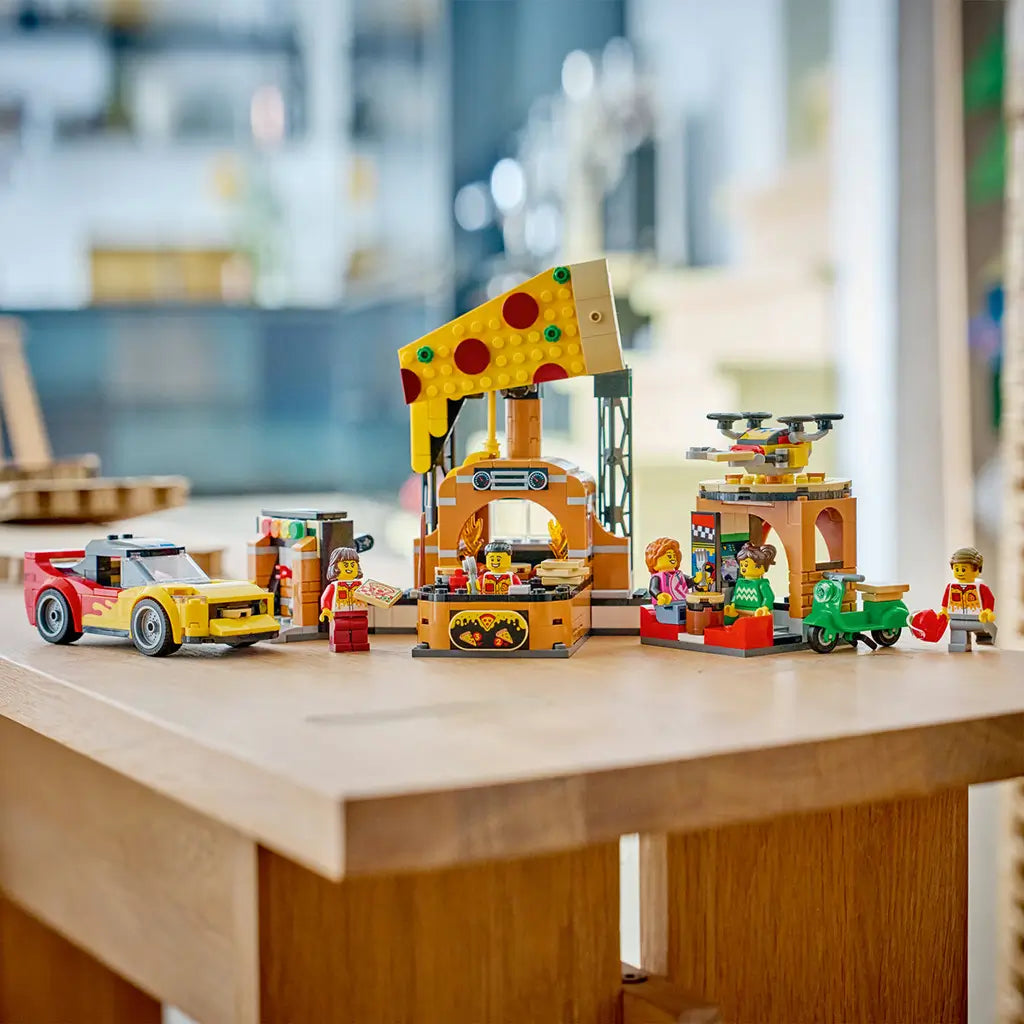 LEGO City Pizza Delivery Experience in yellow, brown, red, and green plastic bricks features a pizza shop, sports car, scooter, drone, and minifigures on a kitchen table.