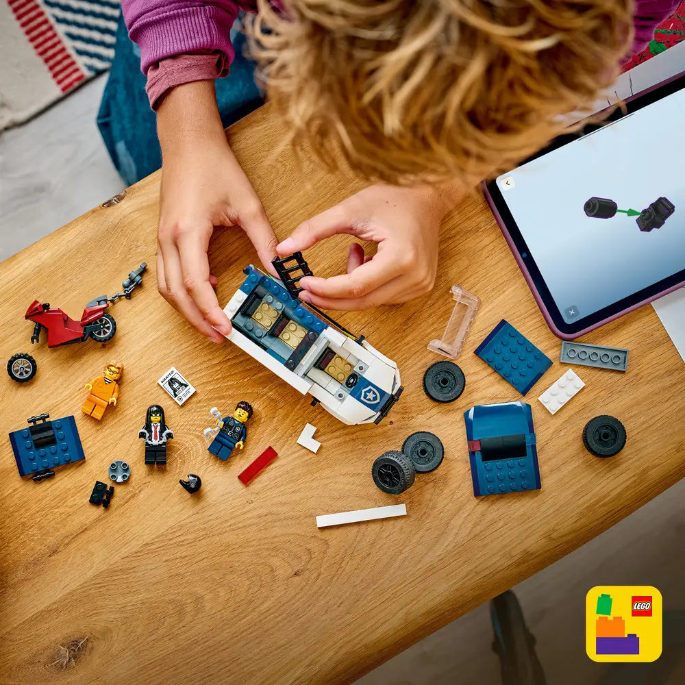 LEGO City police prisoner transport van building scene showing dark blue and white plastic pieces, wheels, minifigures, and accessories arranged on a wooden table as a child follows step by step digital instructions on a tablet.