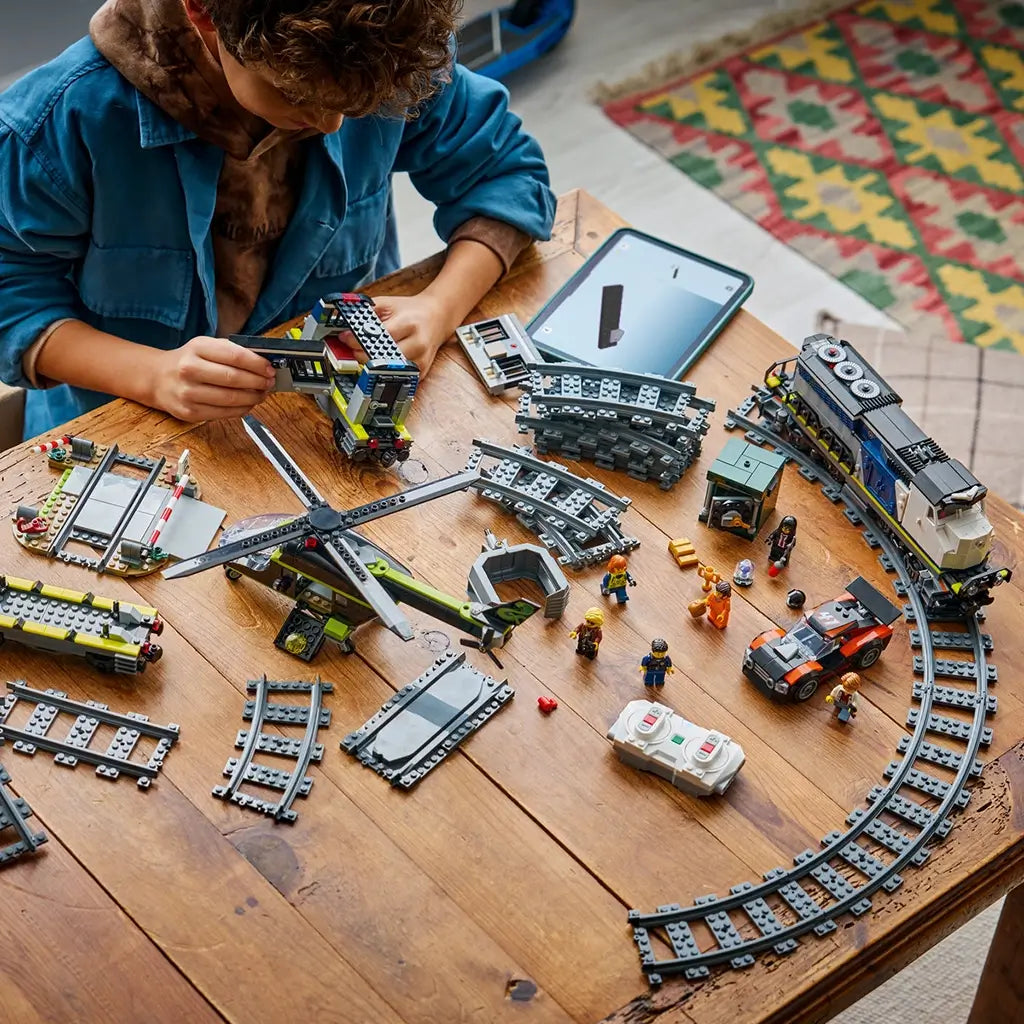 LEGO City construction play in blue and gray plastic pieces, train cars, helicopter, and track parts, building toy in progress on a wooden table with tools and figures nearby.