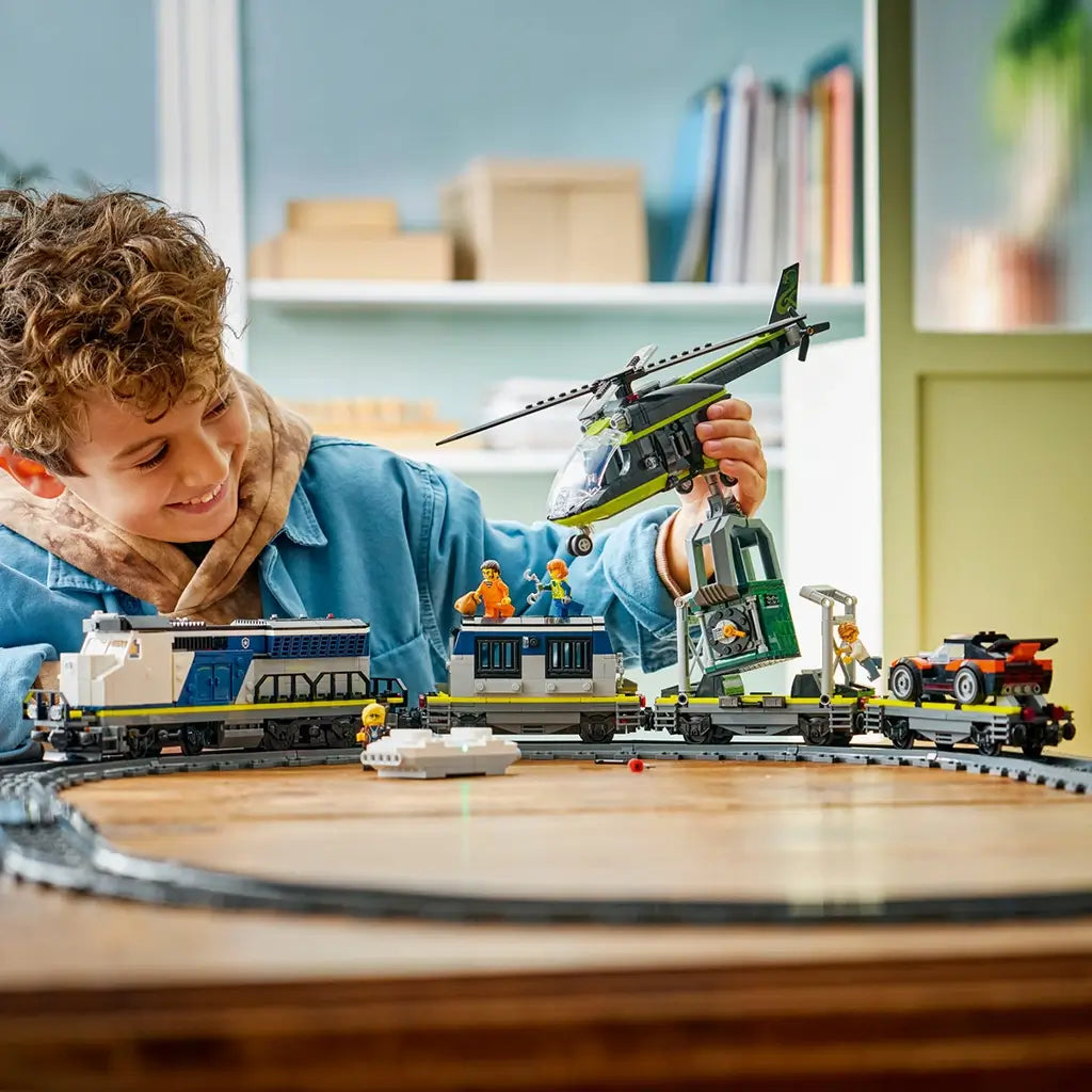 LEGO City train toy in blue, gray, and black plastic, long connected cars with helicopter and figures, action play scene with a child interacting at a wooden table indoor setting.