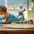 LEGO City train toy in blue, gray, and black plastic, long connected cars with helicopter and figures, action play scene with a child interacting at a wooden table indoor setting.