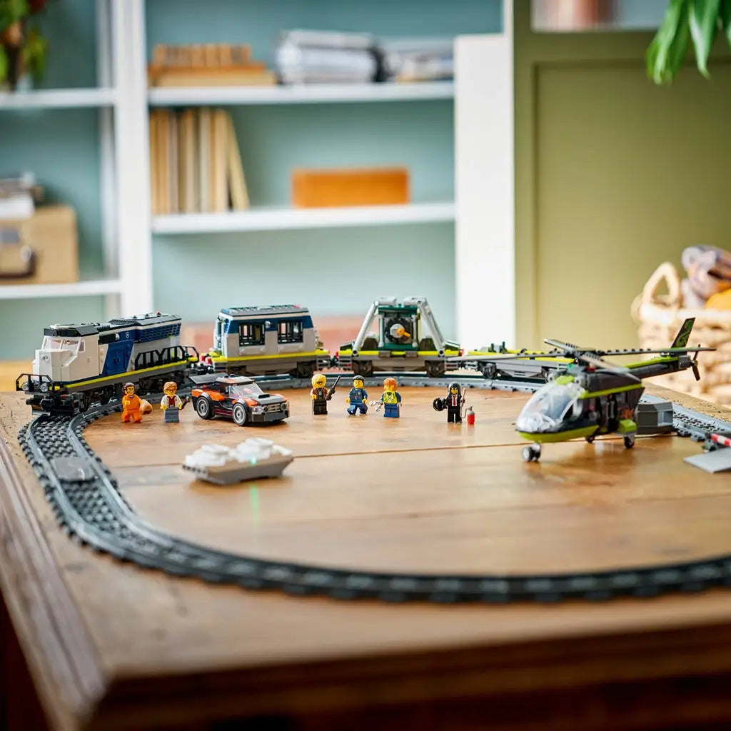 LEGO City train set display in blue and gray plastic, long track with locomotive, cargo cars, helicopter, and figures, arranged toy scene on a wooden table with home interior background.