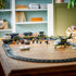 LEGO City train set display in blue and gray plastic, long track with locomotive, cargo cars, helicopter, and figures, arranged toy scene on a wooden table with home interior background.