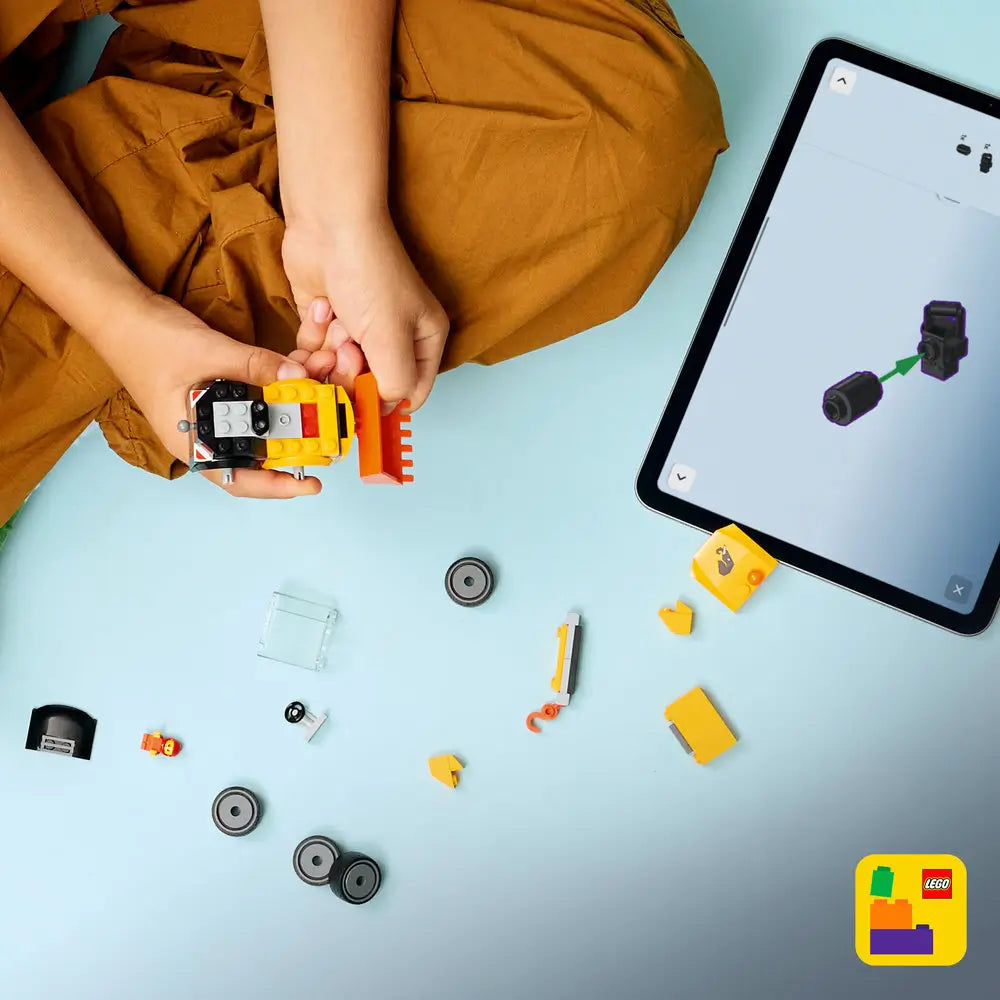 LEGO City Loader building activity showing yellow and black plastic pieces, wheels, bucket parts, and small accessories, assembled by a child beside a tablet displaying step-by-step digital instructions on a light blue surface.