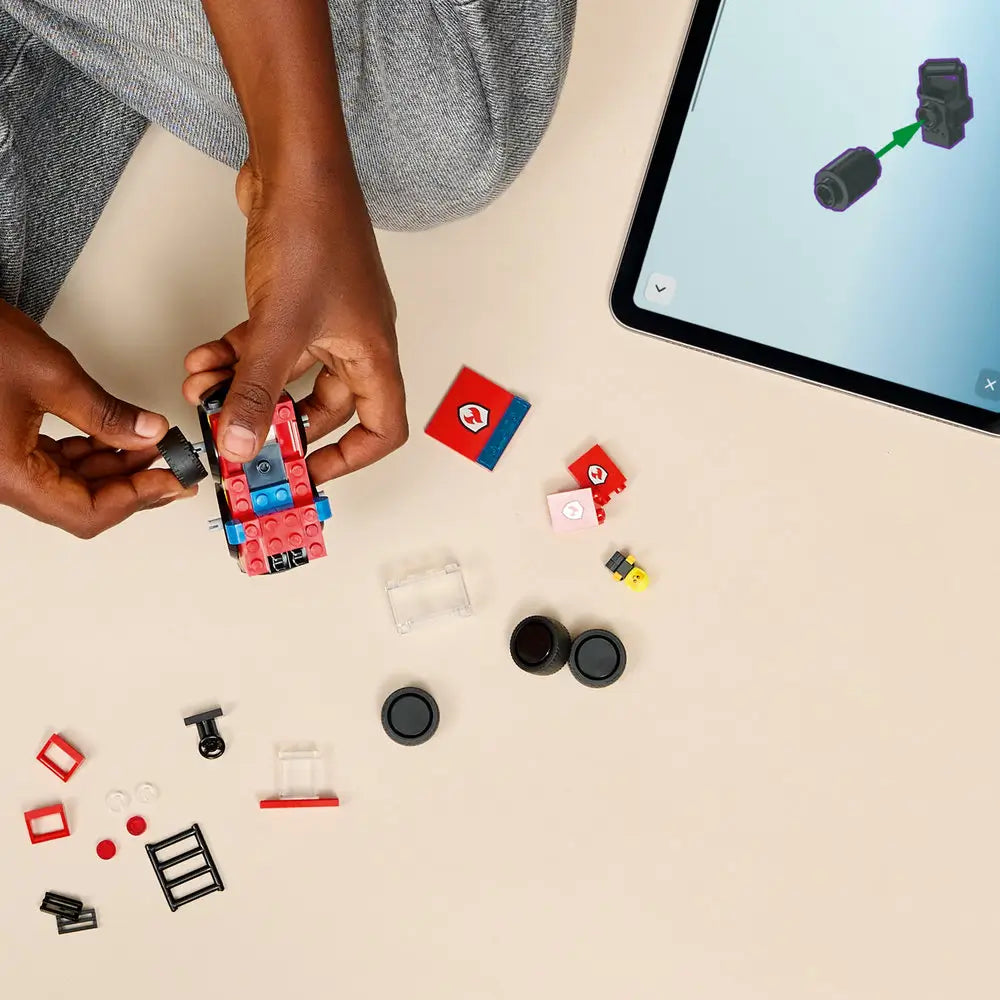LEGO City Fire Truck building activity showing red and black plastic pieces, wheels, ladder parts, and small accessories, assembled by a child beside a tablet displaying step-by-step digital instructions on a light surface.