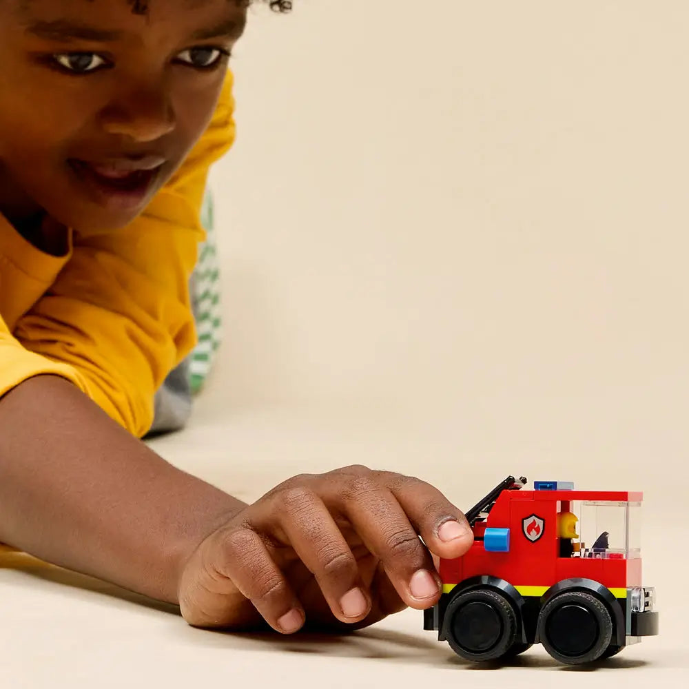 LEGO City Fire Truck lifestyle scene showing a child pushing the completed red fire truck toy, clear cab, ladder accessory, six-wheel design, and firefighter minifigure on a smooth tabletop background.