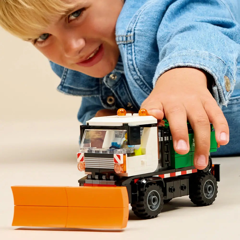 LEGO City snowplow toy played with by a child, showing hands pushing a white and green truck with an orange plow blade, rolling wheels, and interactive winter play features.