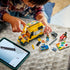 LEGO City delivery van building set shown during assembly, featuring a yellow plastic van body, loose bricks, minifigures, accessories, and a tablet displaying digital building instructions on a table.