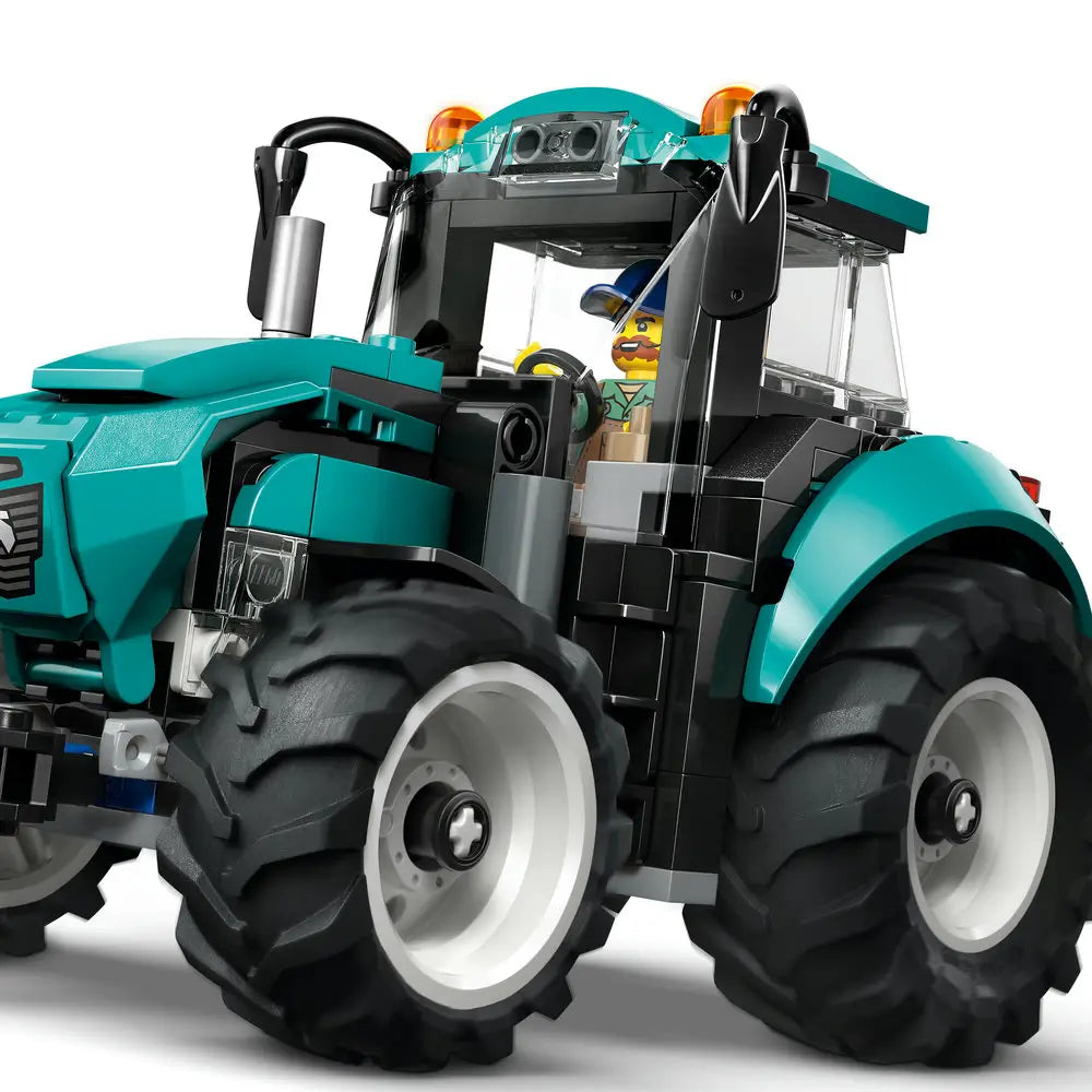 LEGO City tractor close-up showing a teal plastic body, oversized black rubber-style tires, clear cab windows, roof lights, and a seated farmer minifigure operating the vehicle.