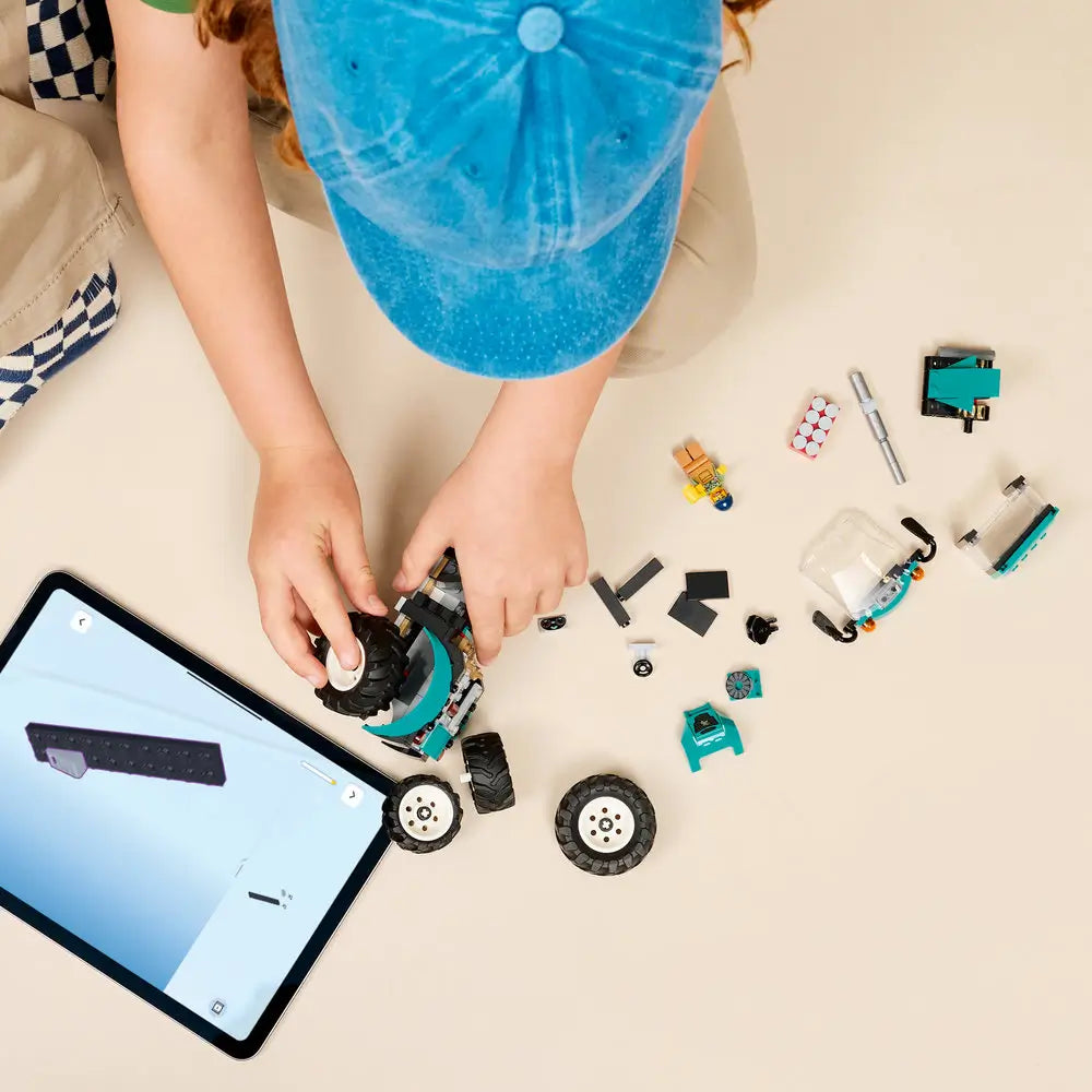 LEGO City tractor building set shown during assembly, featuring teal and black plastic tractor parts, loose wheels, minifigure pieces, and a tablet displaying digital instructions on a tabletop.