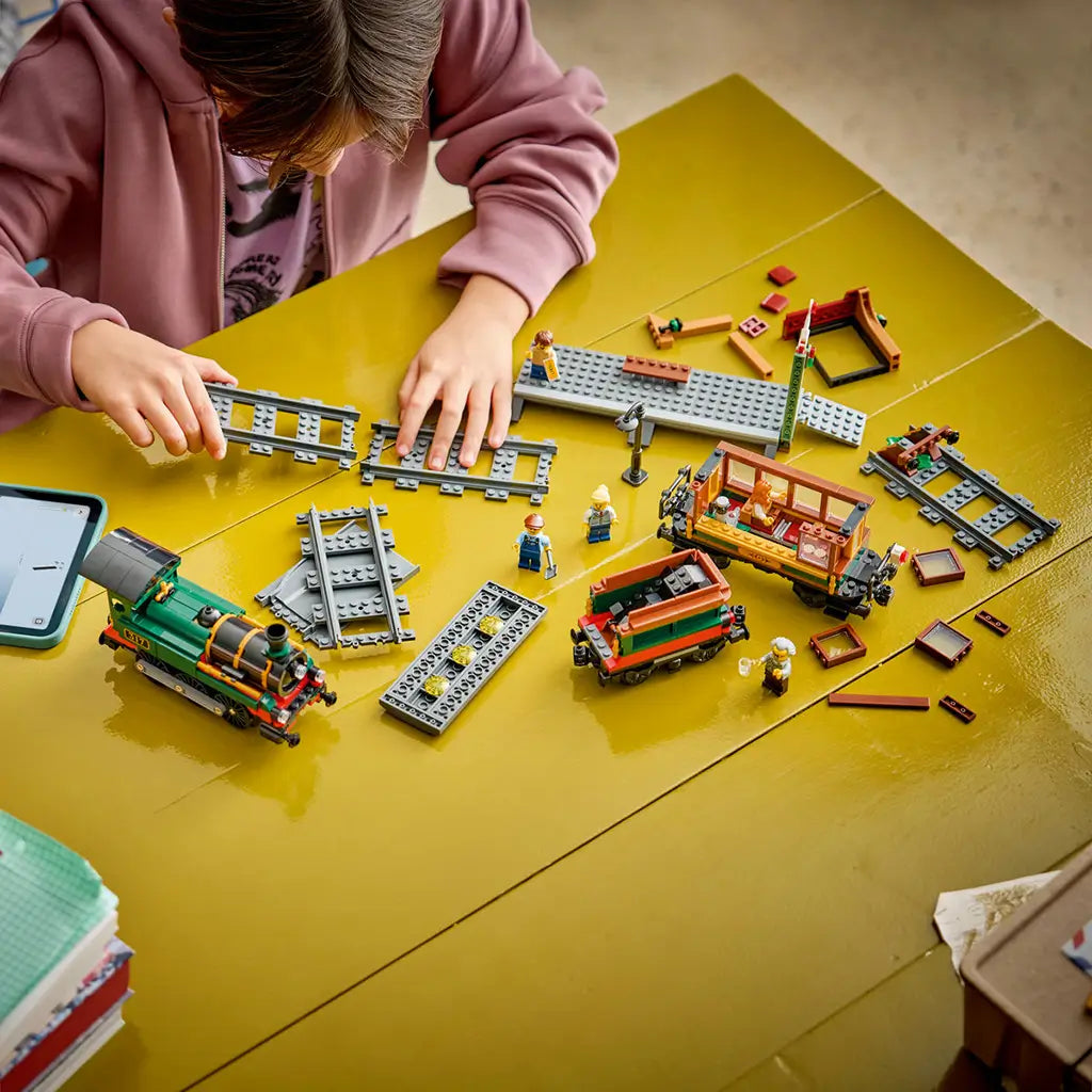 LEGO City Vintage Steam Train in green, black, red, brown, and gray plastic bricks is assembled by a child with track sections, cars, minifigures, and a tablet on a table.