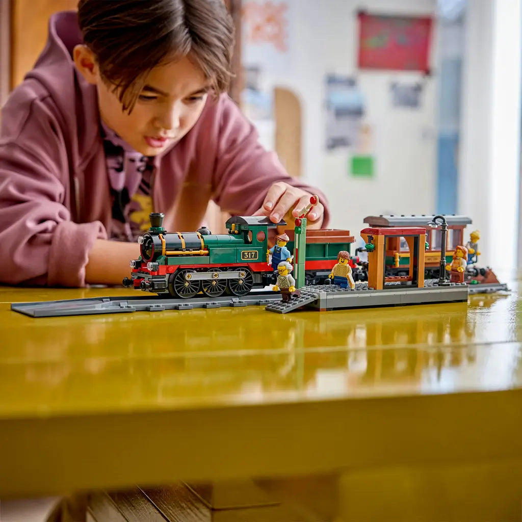 LEGO City Vintage Steam Train in green, black, red, brown, and gray plastic bricks sits on track as a child plays with the locomotive, passenger car, station, and minifigures.