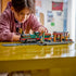 LEGO City Vintage Steam Train in green, black, red, brown, and gray plastic bricks sits on track as a child plays with the locomotive, passenger car, station, and minifigures.