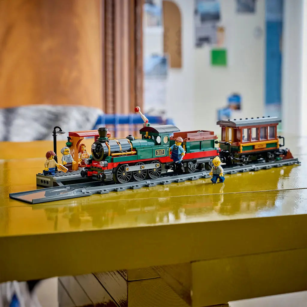 LEGO City Vintage Steam Train in green, black, red, and brown plastic bricks sits on gray track with a passenger car, station platform, and minifigures on a yellow table.