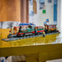 LEGO City Vintage Steam Train in green, black, red, and brown plastic bricks sits on gray track with a passenger car, station platform, and minifigures on a yellow table.