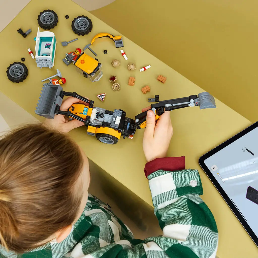 LEGO City Backhoe Loader building toy in yellow and black plastic is assembled by a child at a table with loose wheels, bricks, minifigures and a digital tablet.