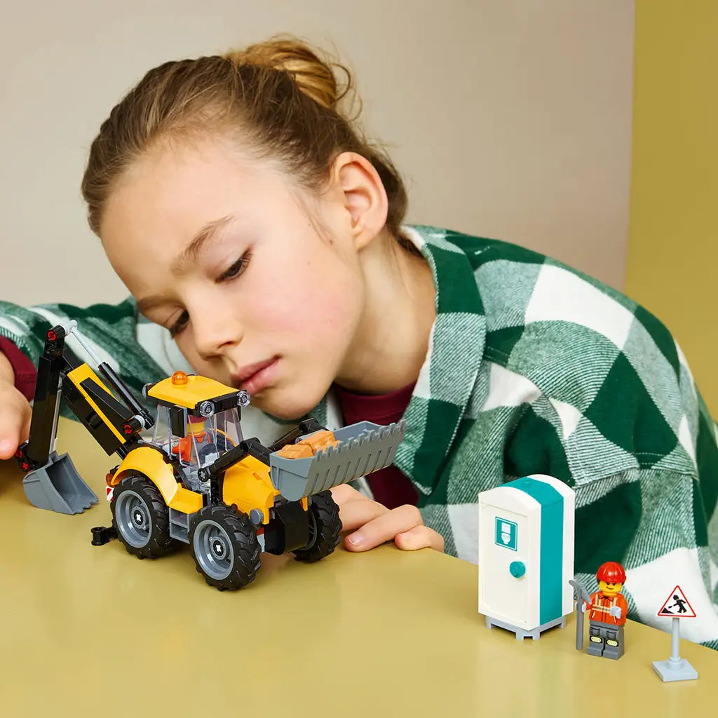 LEGO City Backhoe Loader toy in yellow and black plastic is operated by a child on a tabletop beside a portable toilet, worker minifigure and construction road sign.