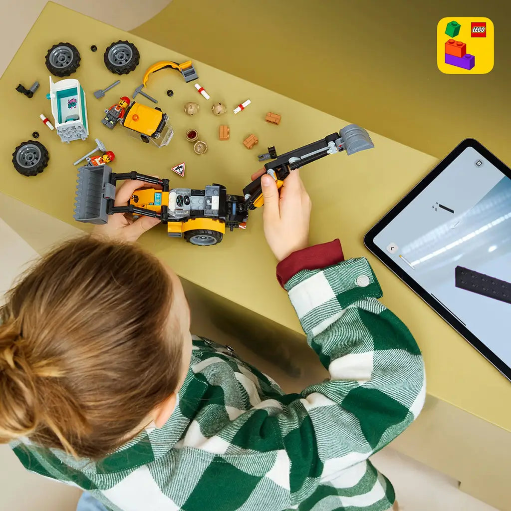 LEGO City Backhoe Loader building toy in yellow and black plastic is assembled by a child beside loose parts and a tablet displaying digital LEGO instructions.