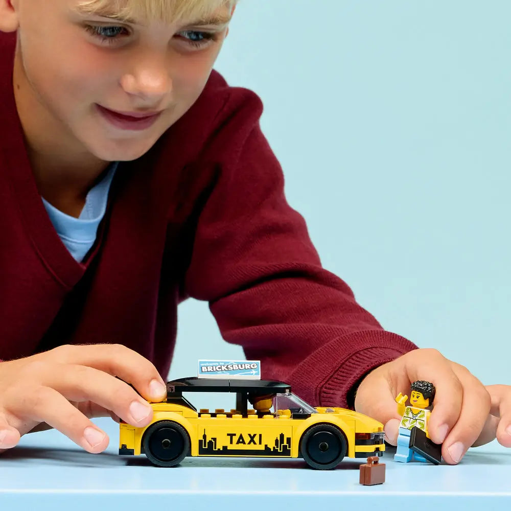 LEGO City taxi play scene with a child holding a yellow brick-built taxi car and minifigure, showing scale, rolling wheels, and imaginative roleplay against a soft light blue studio background.
