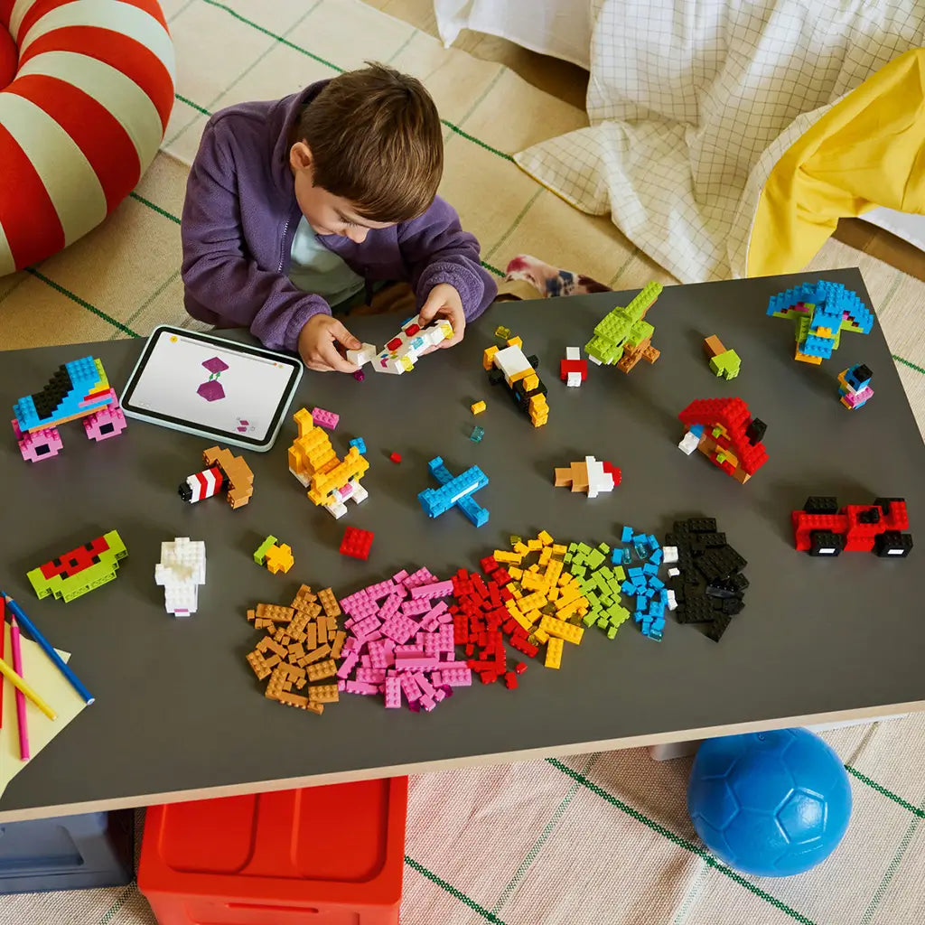 LEGO Classic colorful bricks in pink, red, yellow, green, blue, black, white, and brown surround a child building a model beside a tablet on a playroom table.