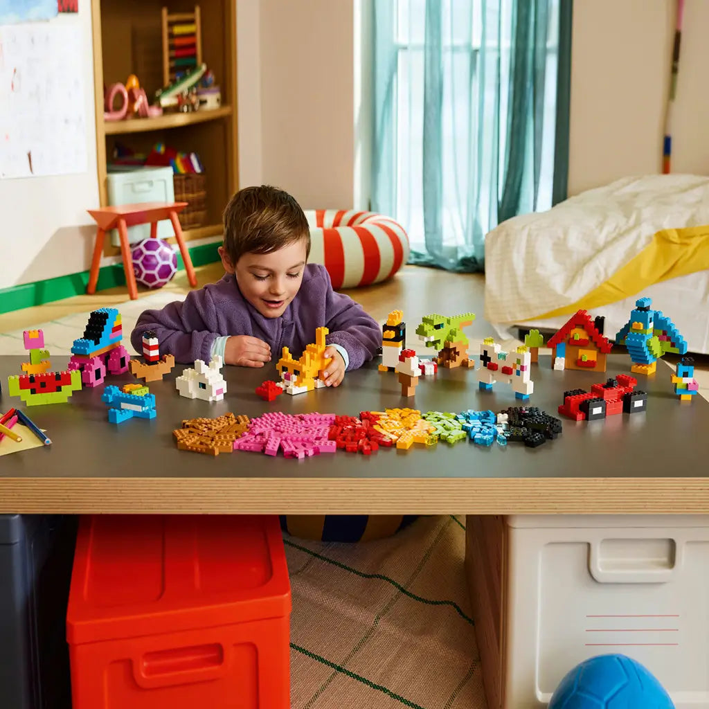 LEGO Classic colorful bricks in bright plastic colors form animals, vehicles, food, and a house as a child builds an orange cat model at a table in a playroom.
