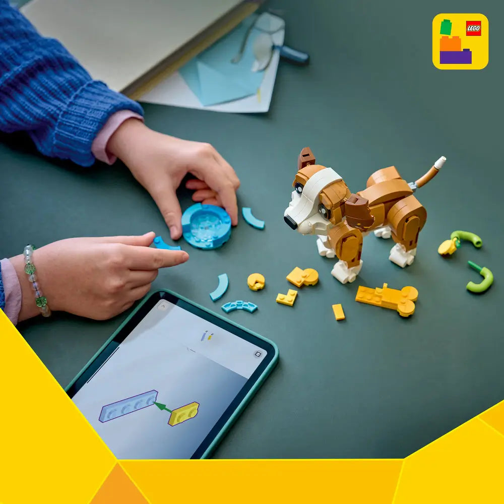 LEGO Creator puppy building set displayed on a green tabletop with scattered colorful plastic bricks, showing a partially built brown and white dog figure beside digital building instructions on a tablet.
