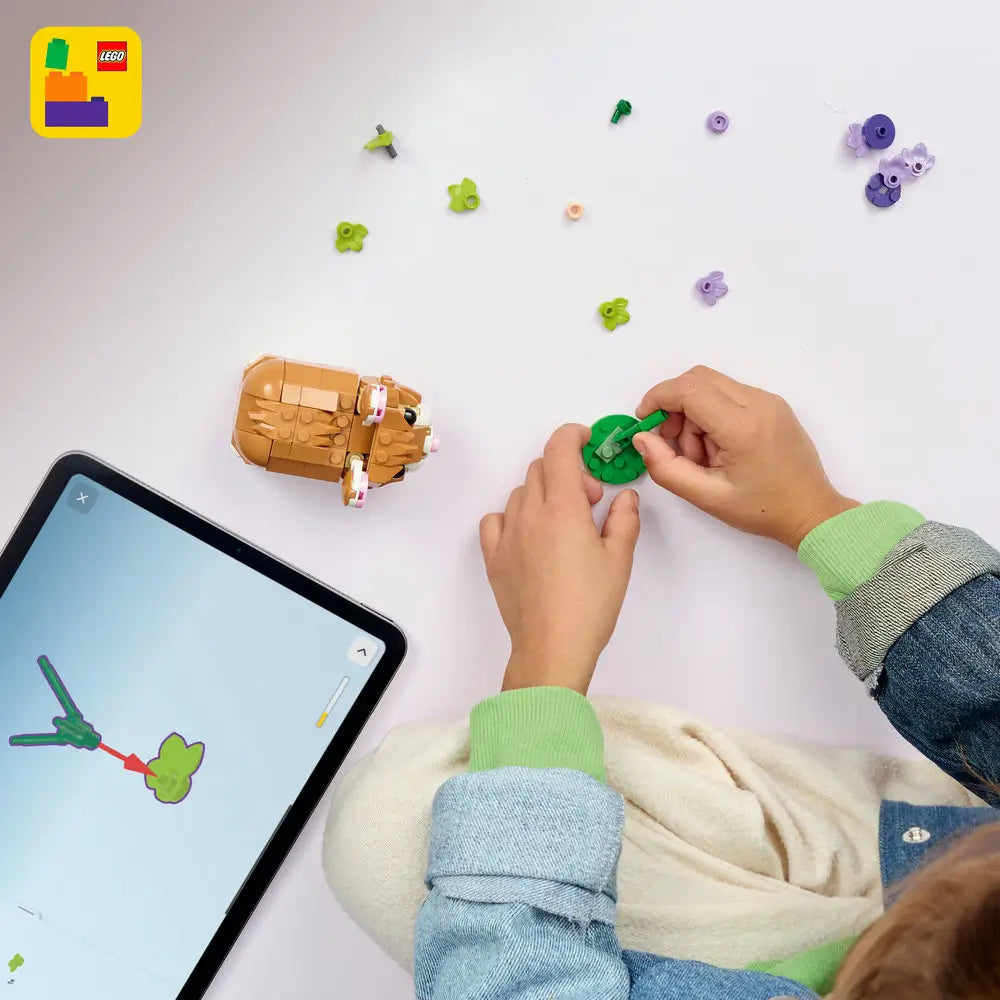 LEGO Creator hamster building set arranged on a white tabletop, showing tan and white plastic bricks, a partially assembled hamster figure, scattered green and purple flower pieces, and a tablet displaying digital step-by-step building instructions.