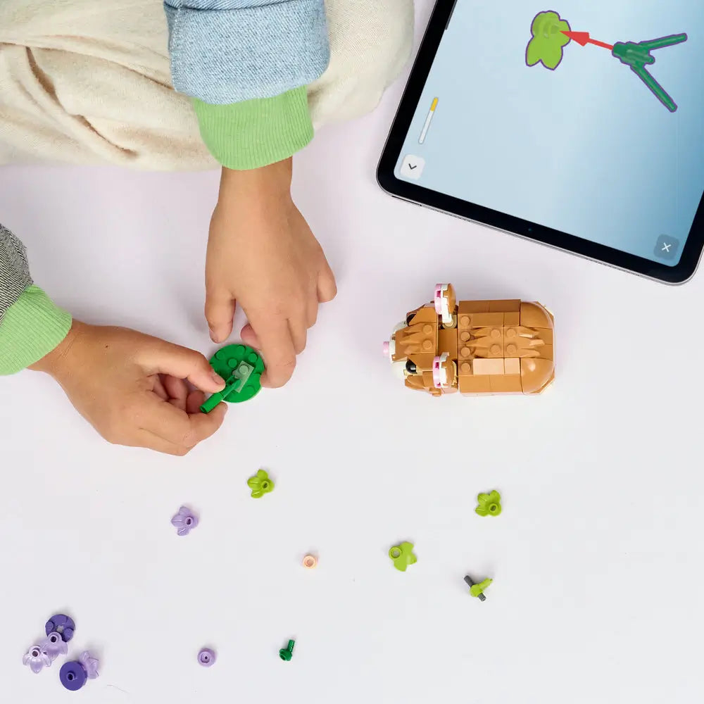 LEGO Creator hamster model being built on a tabletop, featuring tan and white plastic bricks, loose flower and leaf elements, and a partially assembled hamster positioned beside a tablet with interactive building instructions.