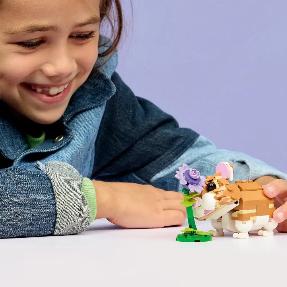 LEGO Creator hamster toy in tan and white plastic bricks as a child plays with the hamster and purple flower, highlighting scale, rounded details, and imaginative indoor animal-themed play.