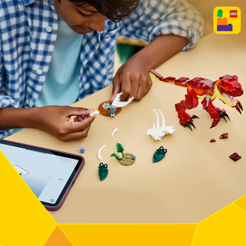 LEGO Creator dinosaur building set arranged on a tabletop, showing red and yellow plastic bricks, a partially assembled dinosaur figure, scattered bone and plant elements, and a tablet displaying digital step-by-step building instructions.