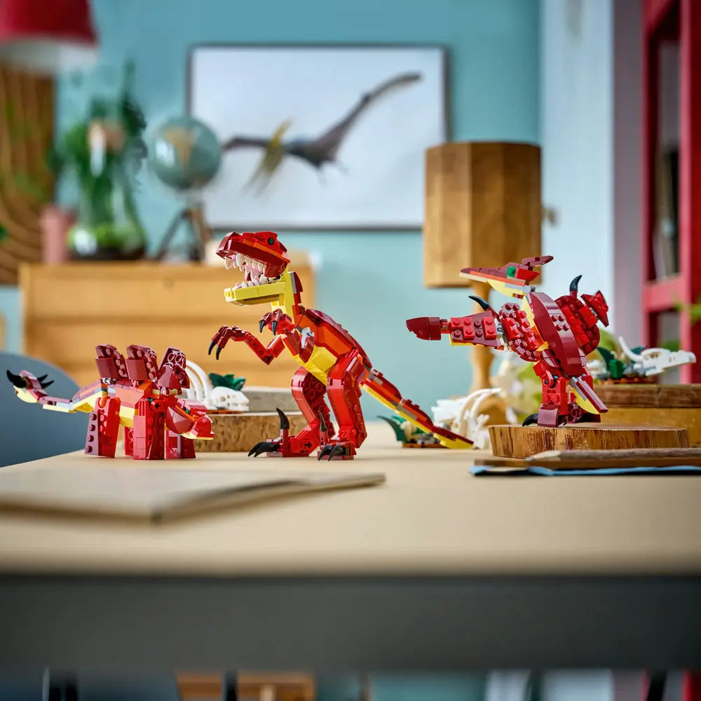 LEGO Creator 3-in-1 dinosaur models displayed together on a tabletop, showing a dinosaur, alternate creature builds, and accessories made from smooth red and yellow plastic bricks in a bright indoor setting.