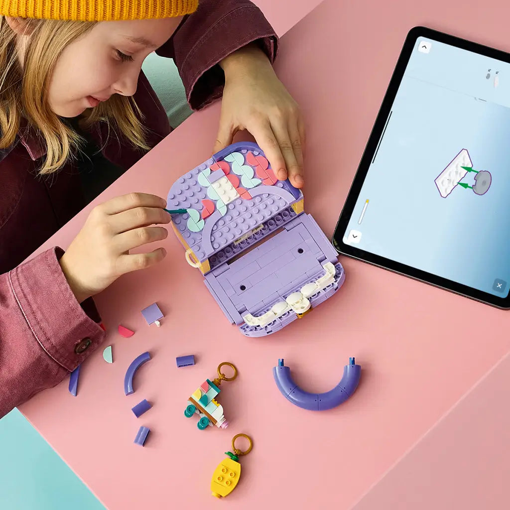 LEGO Creator Fashion Bag in lavender, purple, coral and mint plastic, as a child assembles the open handbag beside loose bricks, charms and digital instructions on a pink table.
