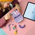 LEGO Creator Fashion Bag in lavender, purple, coral and mint plastic, as a child assembles the open handbag beside loose bricks, charms and digital instructions on a pink table.