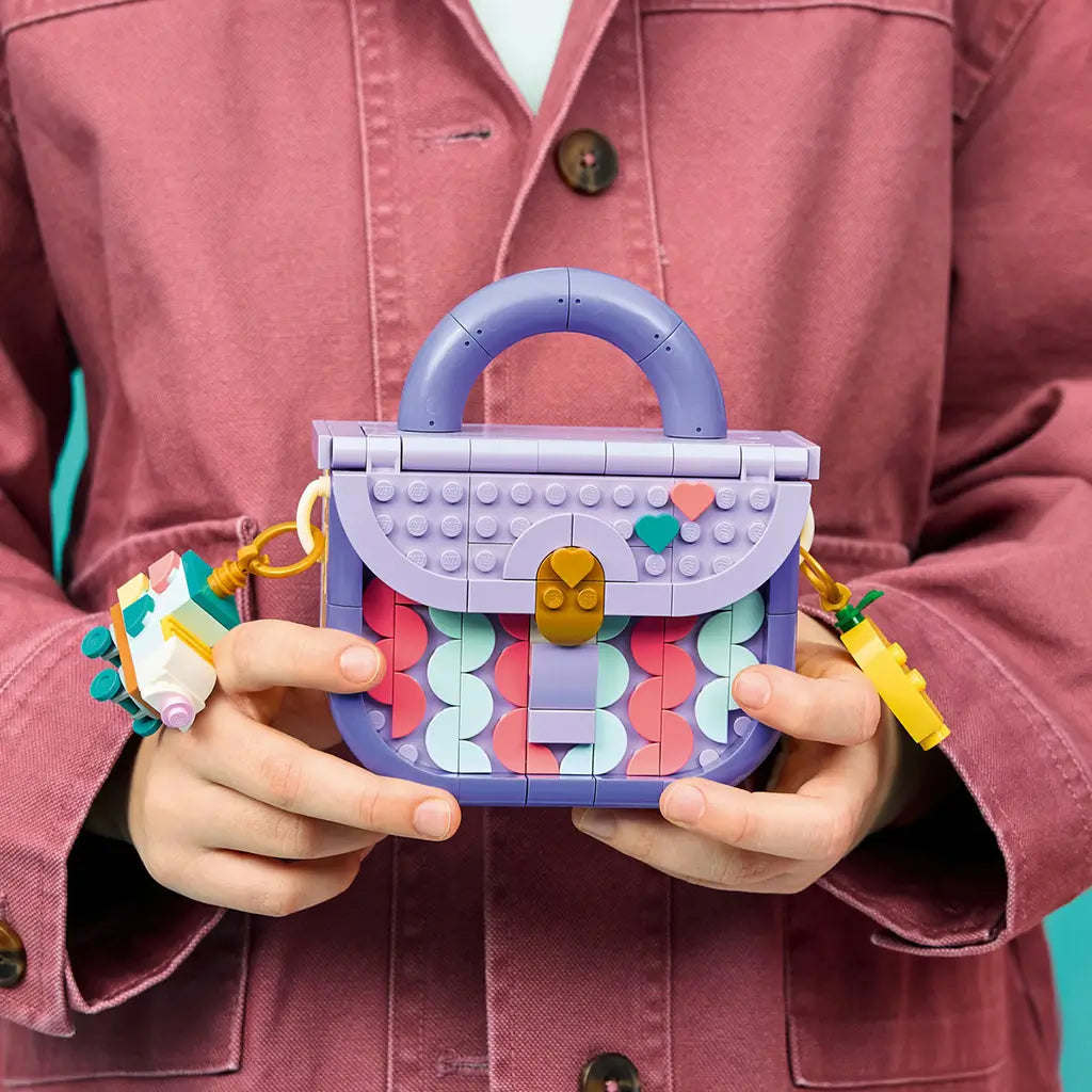 LEGO Creator Fashion Bag in lavender, purple, coral and mint plastic, held by a child to show its rounded handle, gold clasp, heart accents, roller skate and yellow key charms.