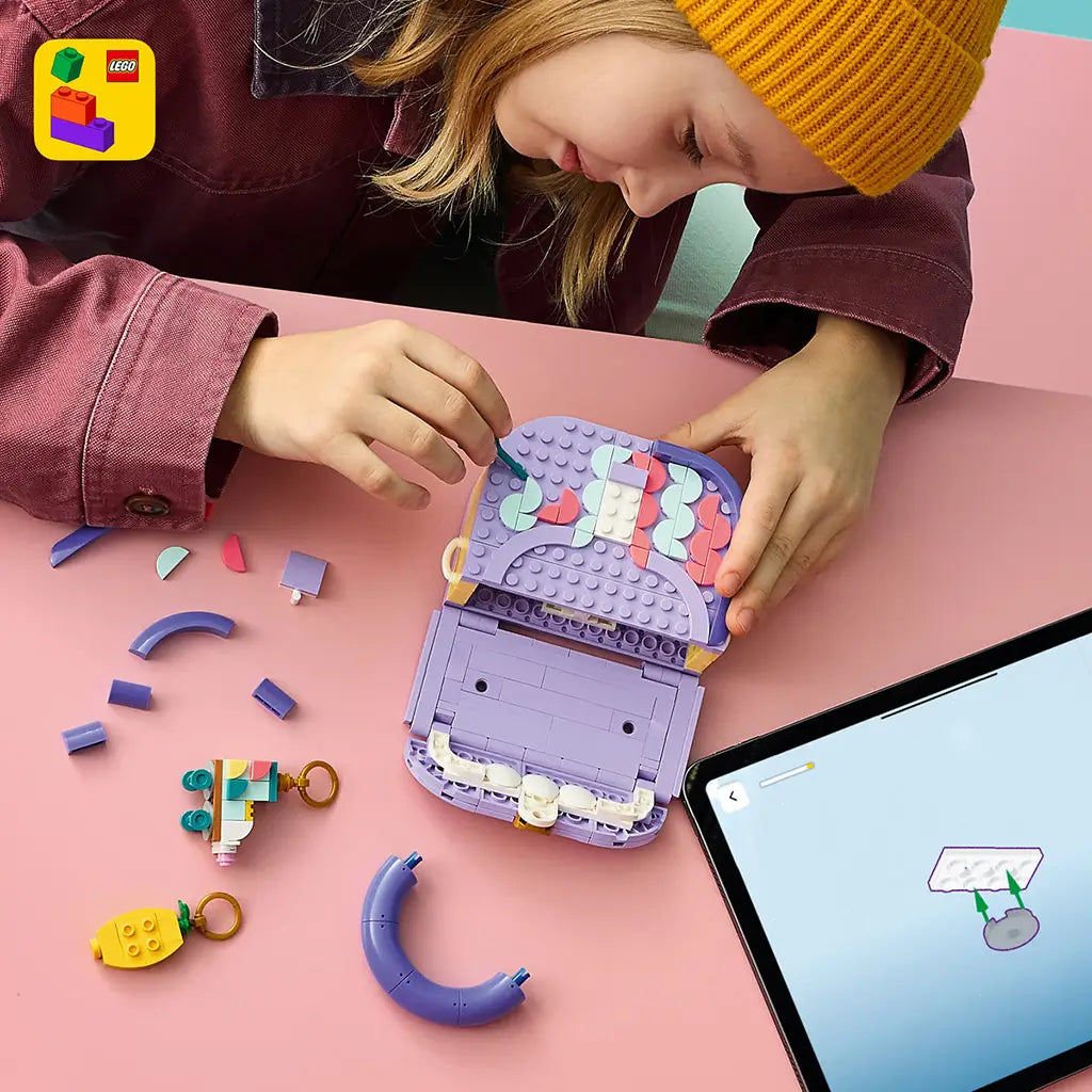 LEGO Creator Fashion Bag in lavender, purple, coral and mint plastic, as a child builds the open handbag among loose pieces, charms and digital instructions on a pink tabletop.