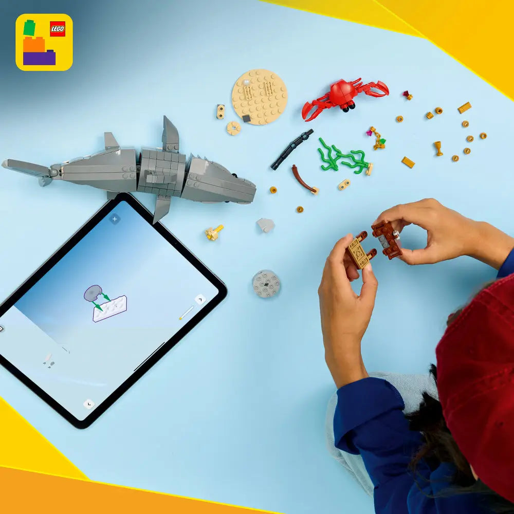 LEGO Creator shark building set arranged on a light blue surface, showing gray plastic bricks, a partially assembled shark model, scattered sea-themed elements, and a tablet displaying digital step-by-step building instructions.