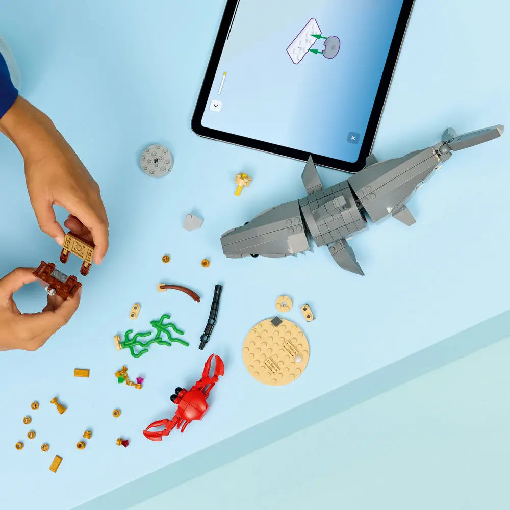 LEGO Creator shark model being built on a tabletop, featuring gray plastic bricks, loose ocean-themed parts, a red crab, a treasure chest, and a tablet nearby showing interactive building instructions.